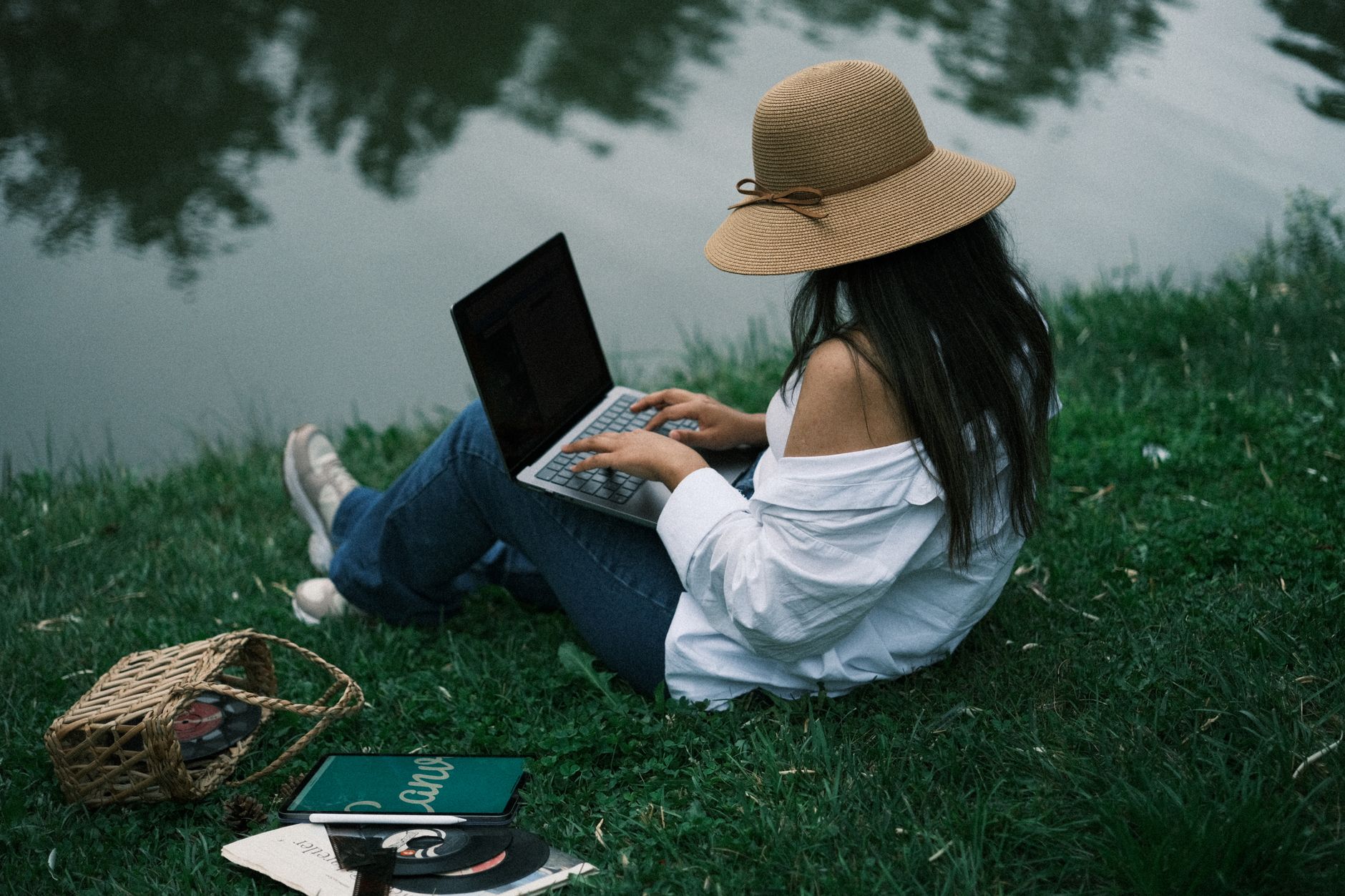 Woman wearing a hat using a laptop while sitting by a tranquil lake in a park.