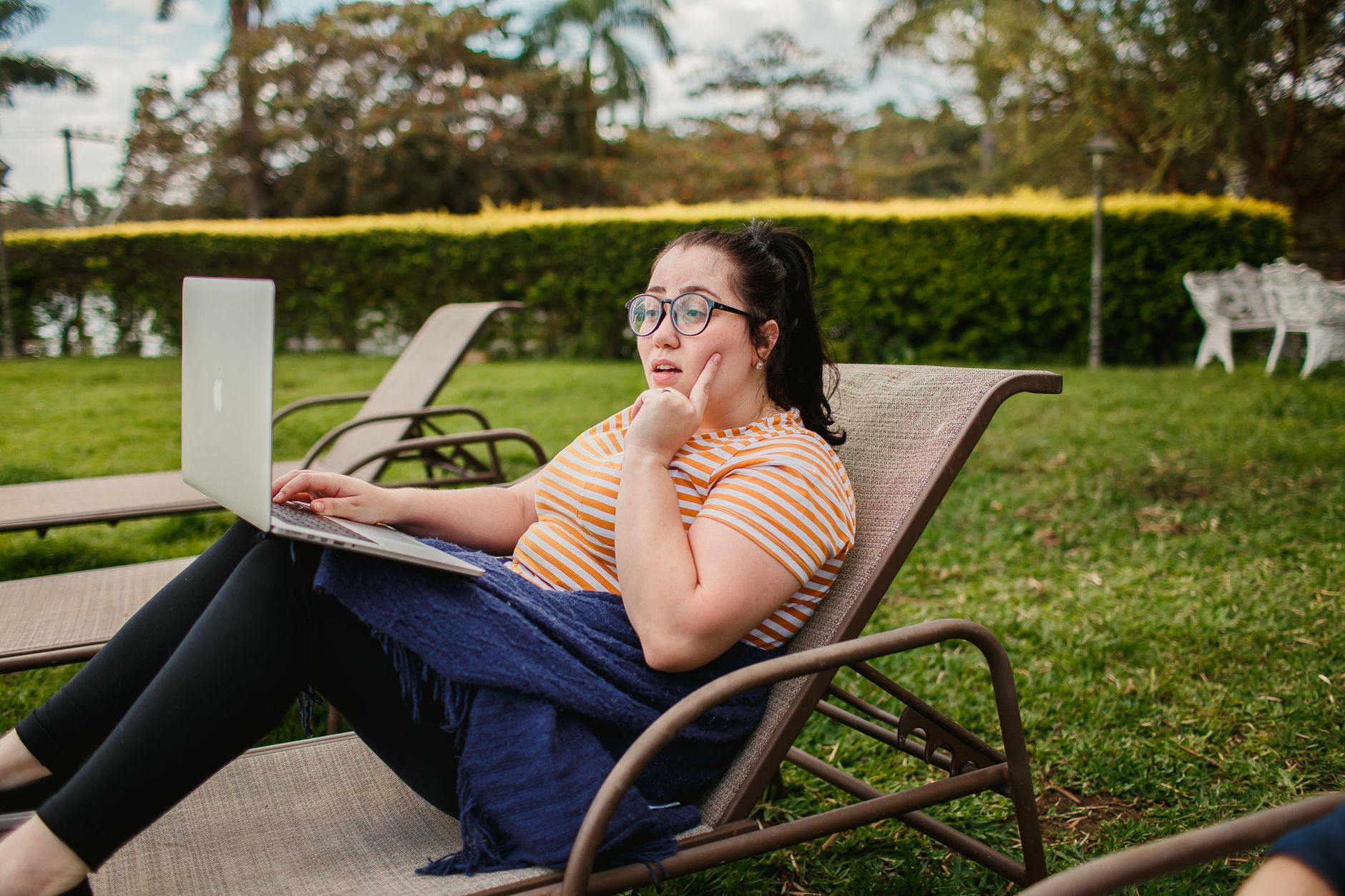 Woman relaxing on a lounge chair with a laptop in a lush green garden, enjoying a sunny day.